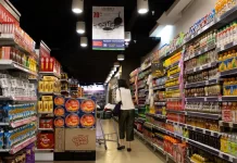 Jak skutecznie komunikować promocje wizualnie w punkcie sprzedaży Kobieta z wózkiem idzie alejką supermarketu pełną produktów i promocji.