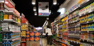 Kobieta z wózkiem idzie alejką supermarketu pełną produktów i promocji.