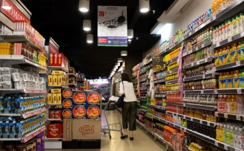 Kobieta z wózkiem idzie alejką supermarketu pełną produktów i promocji.