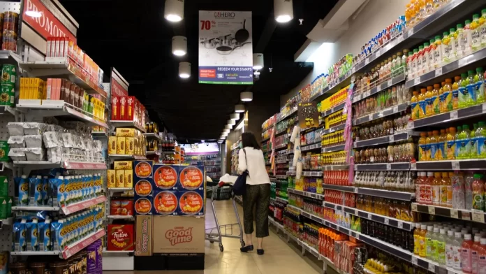 Kobieta z wózkiem idzie alejką supermarketu pełną produktów i promocji.